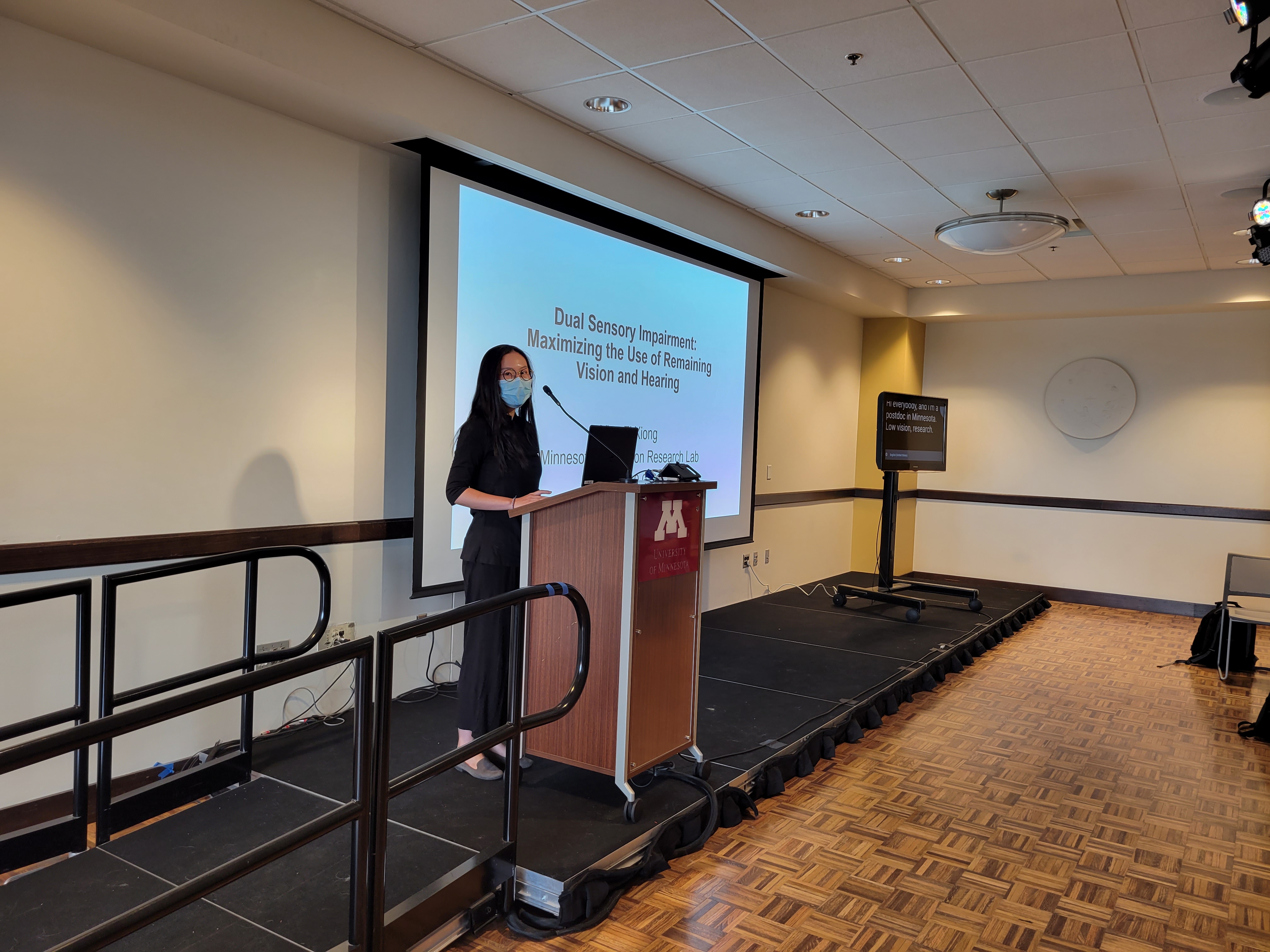 Ying-Zi wearing all black, standing at a podium with her slide presentation behind her on her right.