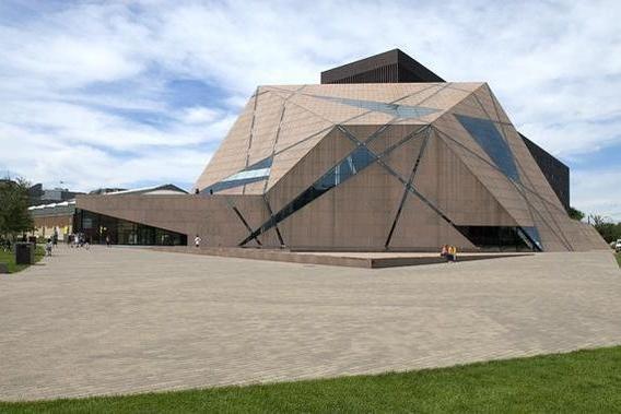 Brown, creatively-shaped building with a patio in front of it. The sky in the background is blue with several clouds appearing.